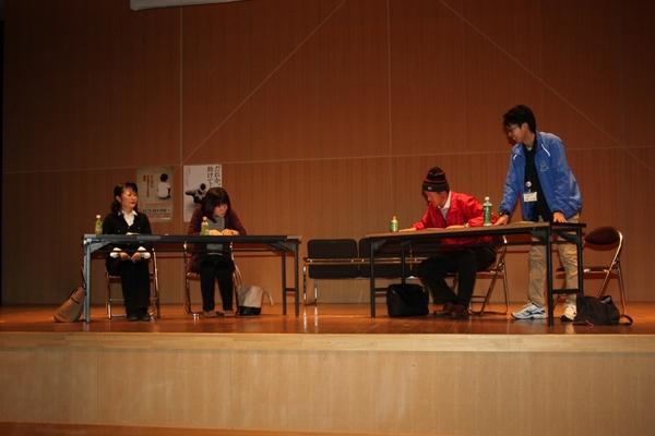 舞台の上で長机と椅子が設置され男女4名で話し合いが行われている写真